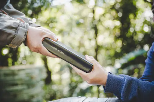 Two hands exchange a Bible outdoors, surrounded by greenery. The moment conveys a sense of sharing and connection in a peaceful setting.