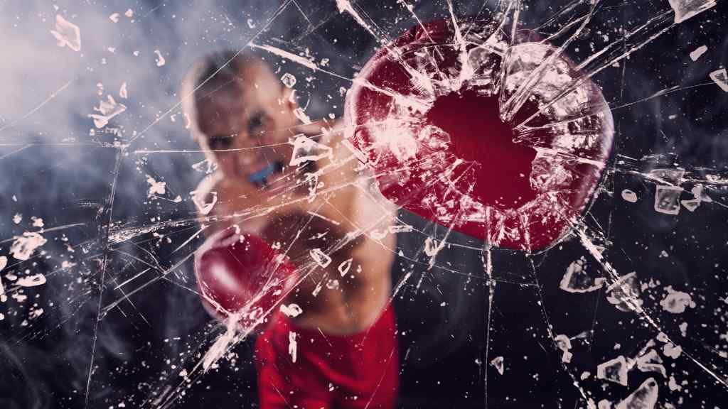 A boxer in red gloves punches at the camera, shattering glass with intense force. Dynamic action and energy are captured against a dark backdrop.