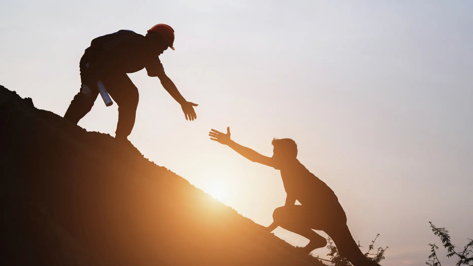 Silhouetted image of two people on a hill at sunset. One helps the other climb, extending hands in a moment of teamwork and support. Warm, motivating tone.