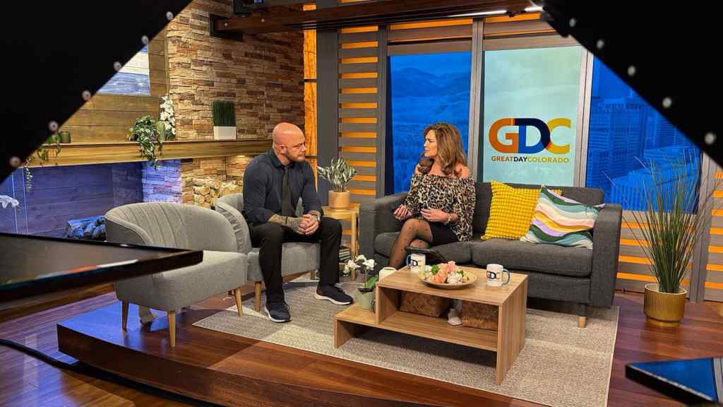 A TV show set with two hosts sitting on sofas. They engage in conversation beside a coffee table with mugs and decor. The backdrop shows "Great Day Colorado."