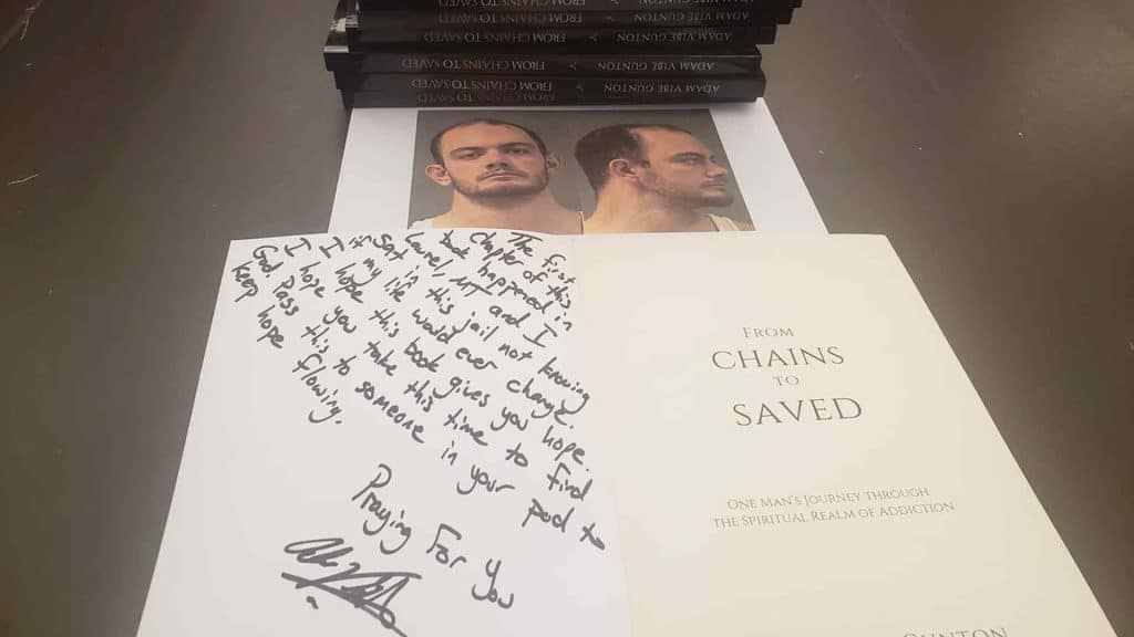 An open book titled "From Chains to Saved" on a table with a handwritten message on one page. Behind it, a stack of similar books and a photo of a man in two angles. The scene conveys a personal and inspirational tone.