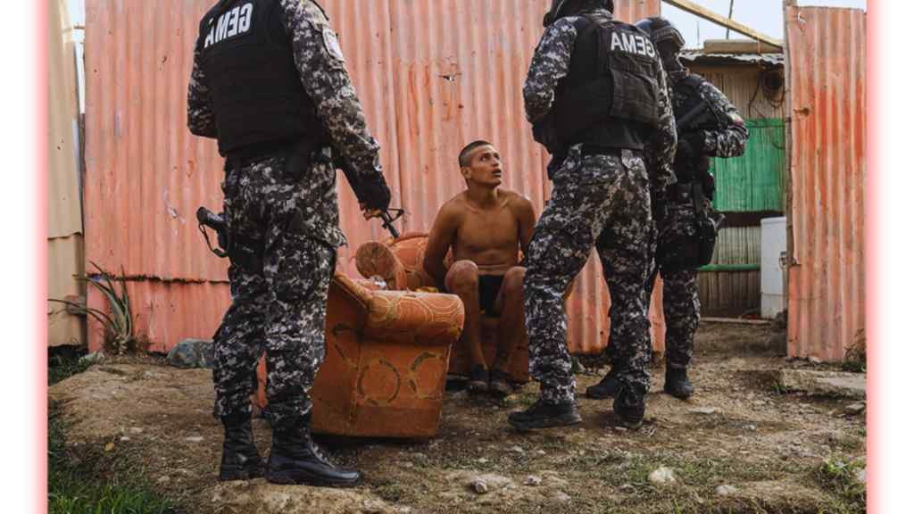 A shirtless man sits on a worn armchair, looking upwards at three armed officers in camouflage uniforms and helmets. The scene is tense, set in a rural area with corrugated metal walls.
