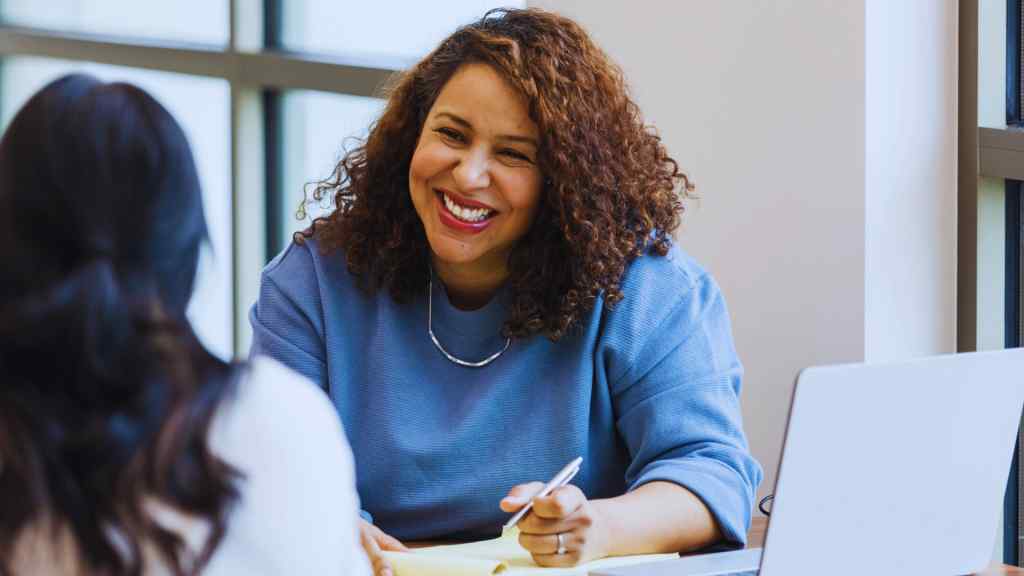 A woman with curly hair, wearing a blue top, smiles warmly during a meeting. She holds a pen over a notebook, sitting at a table with a laptop.