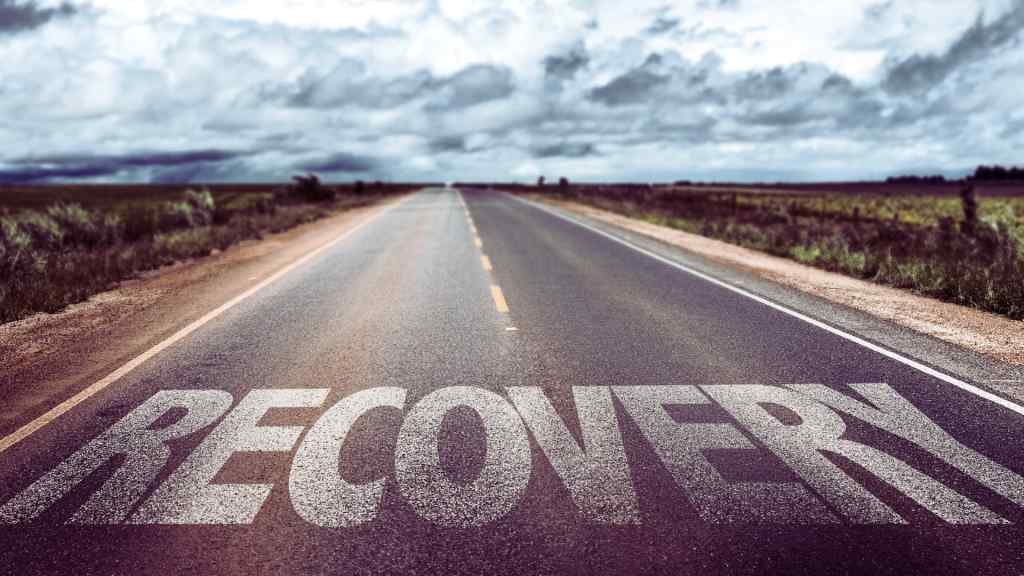 A long, empty highway stretches into the distance under a cloudy sky. The word "RECOVERY" is boldly painted on the road, symbolizing a journey or path forward.