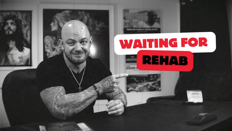 A bald, tattooed man with a friendly expression is seated, pointing to a sign that says "Waiting for Rehab." The setting has framed art on the wall.