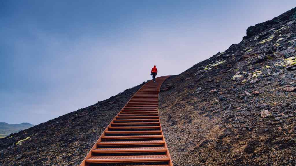 A person in a red jacket walks up a long wooden staircase on a barren hill under a cloudy sky, conveying a sense of solitude and determination.