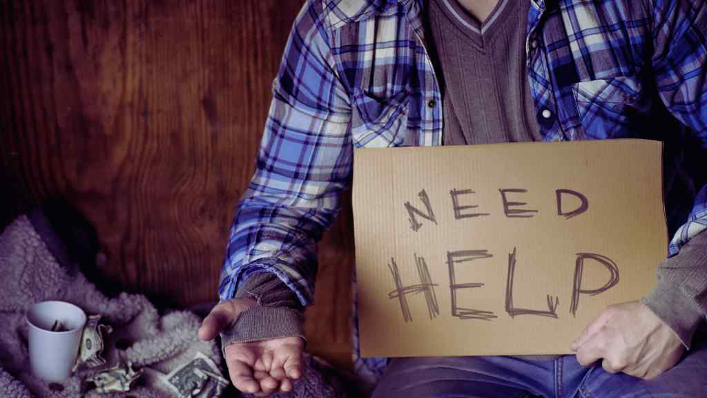 A person in a plaid shirt holds a cardboard sign reading "Need Help." An empty hand is extended, and crumpled dollar bills are visible in a cup nearby. The scene conveys a sense of struggle and plea for assistance.
