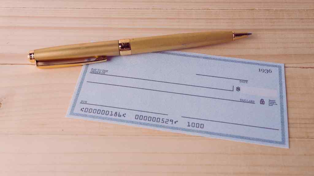 A blank blue check lies on a light wooden surface, with a gold pen placed diagonally across it. The scene conveys readiness for writing.