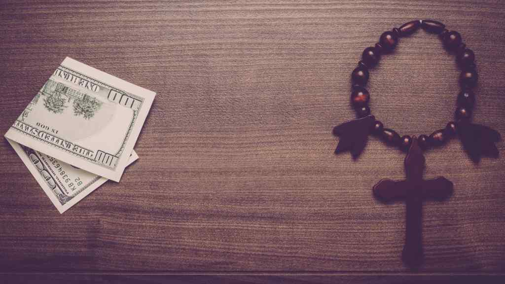 Wooden rosary with cross next to a stack of hundred-dollar bills on a wooden table, conveying a contrast between faith and wealth.