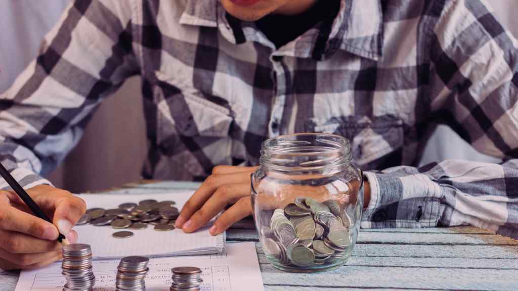 A person in a plaid shirt counts and stacks coins next to a jar filled with more coins, on a table with papers. The scene conveys focus and financial planning.