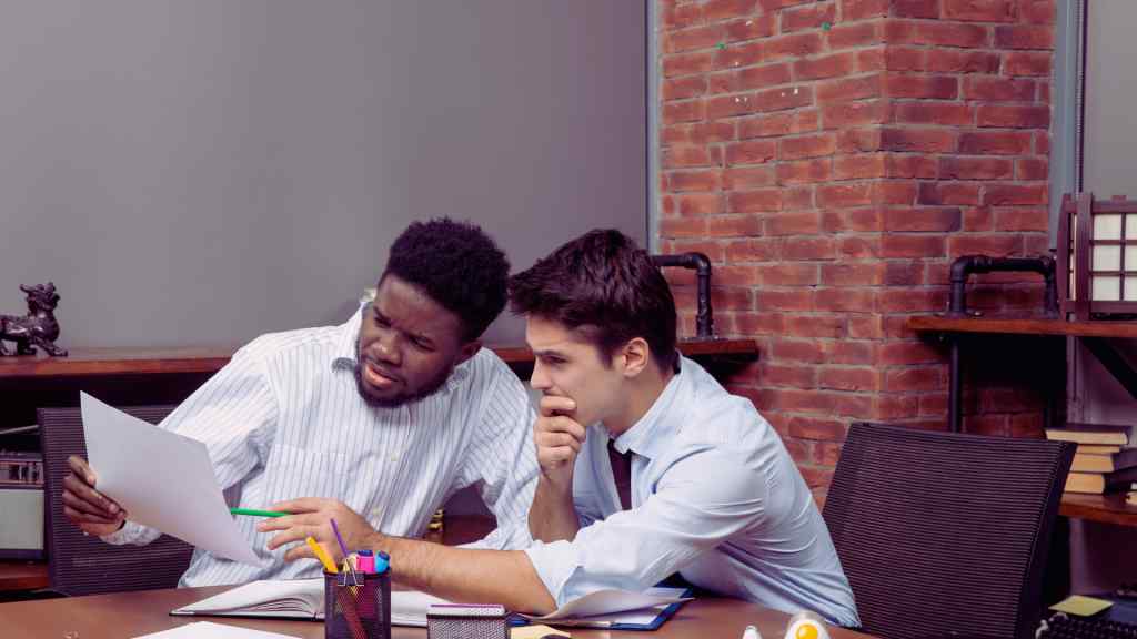 Two men in a modern office study documents intently. One man is pointing with a pen, the other looks thoughtful. Exposed brick wall in the background.