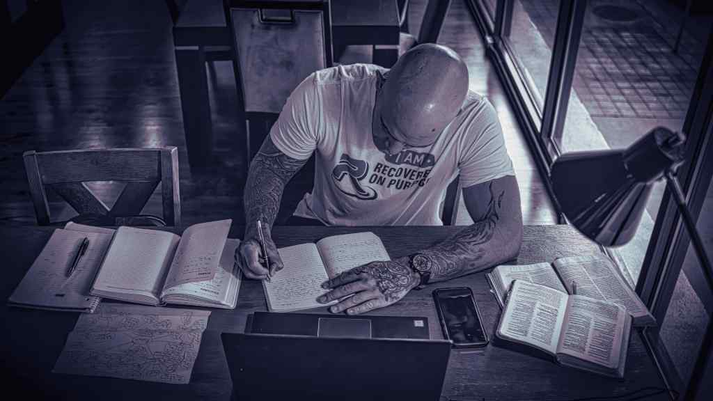 A man with tattoos intensely writes in a notebook at a wooden table, surrounded by open books, a laptop, and a phone. The setting is dimly lit, creating a focused and studious atmosphere.