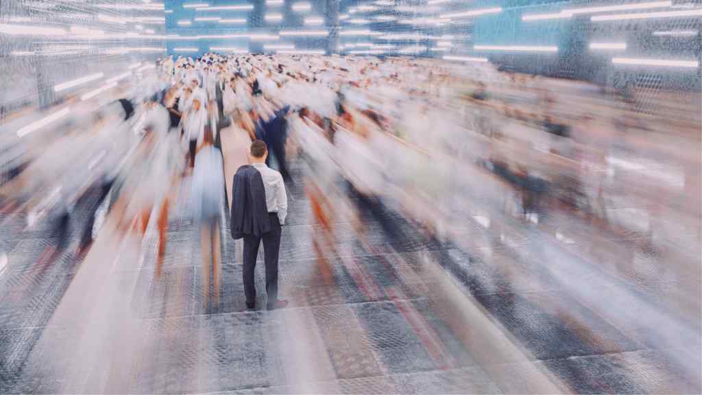 A man stands still in a crowded space, facing a blur of people and lights. The image conveys a sense of isolation amidst chaos and movement.
