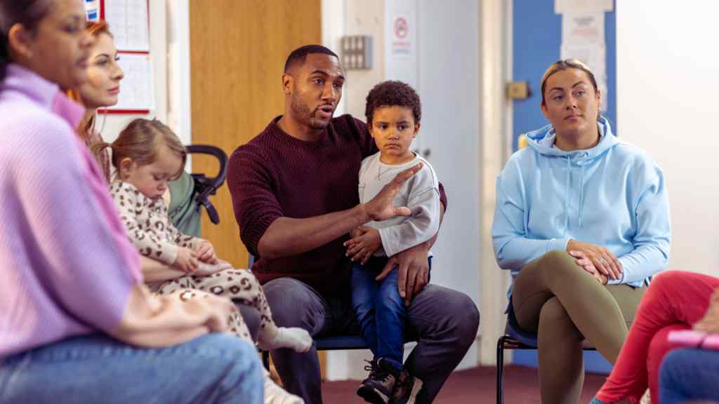 A diverse group of adults and children sit in a circle in a community setting. A man holds a child and speaks, while others listen attentively. The mood is engaged and supportive.