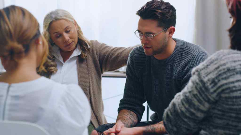 A supportive group setting with three women and a man seated in a circle. The woman in beige comforts the man, conveying empathy and encouragement.