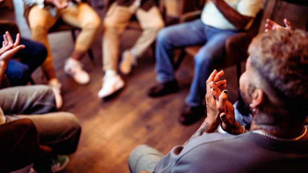 A diverse group of people sit in a circle, gesturing with hands in an engaging discussion. The atmosphere is warm, inviting, and supportive.
