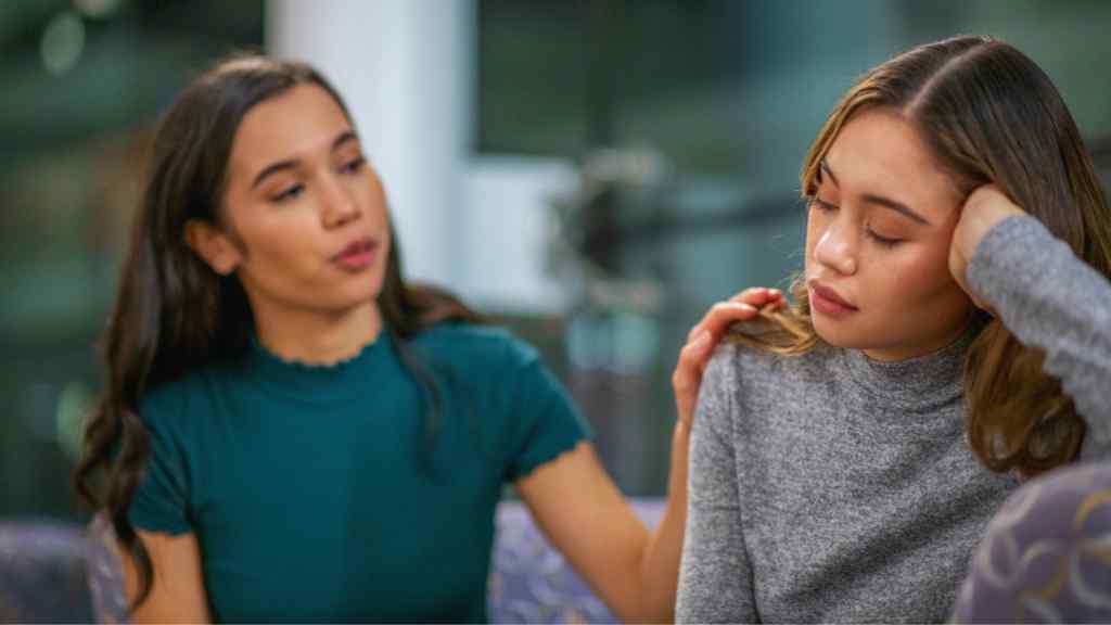 A woman in a teal shirt consoles a sad friend in a gray sweater by gently touching her shoulder. The mood is empathetic and supportive in a cozy indoor setting.