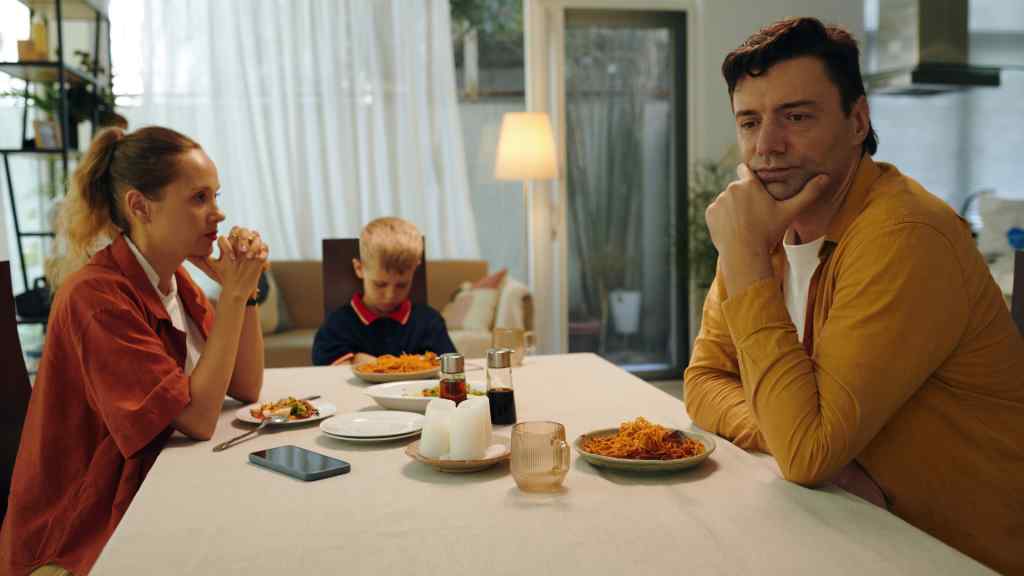 A family of three at a dinner table, appearing thoughtful. The woman and child face the man, who's resting his chin on his hand. Warm lighting.