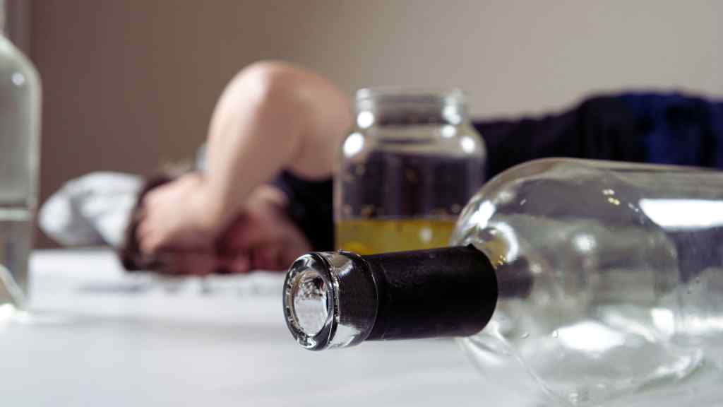 A person lies on a white surface with their arm over their head, surrounded by empty bottles. The scene conveys a sense of exhaustion or hangover.