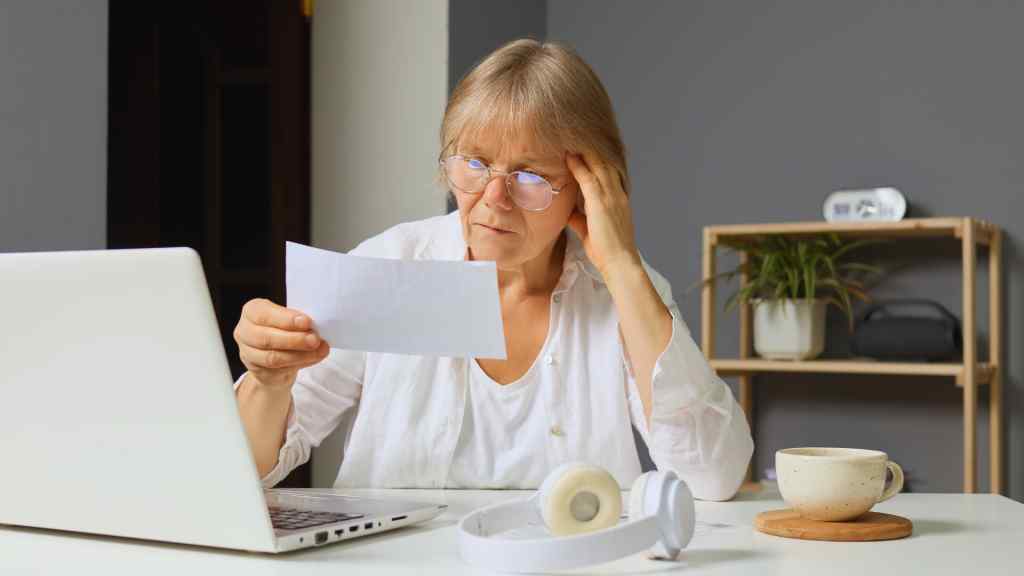 An elderly woman with glasses looks concerned while reading a document at a desk. A laptop, headphones, and a cup are nearby, suggesting a home office setting.