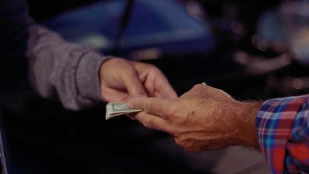 Close-up of two hands exchanging cash in dim lighting. One person wears a gray sweater; the other has a plaid shirt, suggesting a discreet transaction.