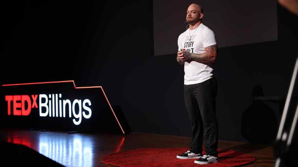A speaker stands on a red circular carpet, delivering a talk at TEDxBillings. He wears a white T-shirt and black pants, conveying confidence and focus.