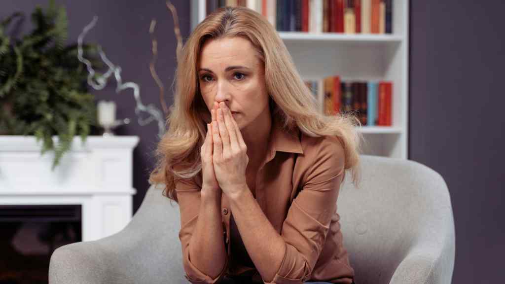A woman with long blonde hair sits pensively in a gray chair, hands clasped near her face, set against a bookshelf and plants, conveying a contemplative mood.