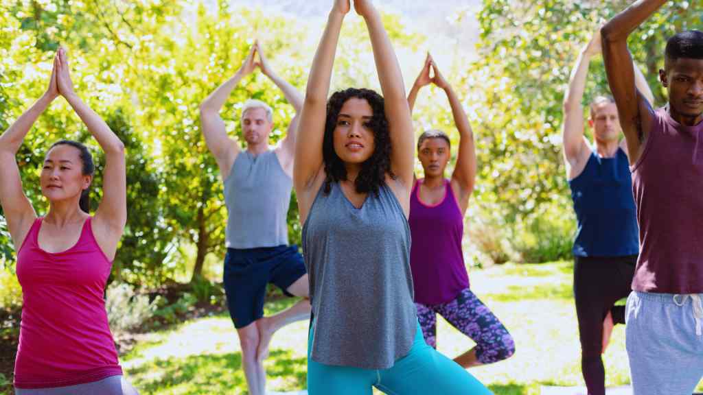 A diverse group of people practice yoga outdoors, standing in a balanced tree pose. Sunlight filters through lush green trees, creating a serene atmosphere.