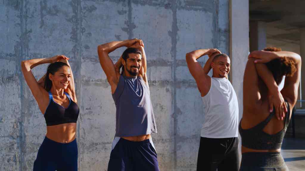 Group of four people stretching outdoors, smiling and dressed in activewear. They appear relaxed and focused, set against a concrete wall background.